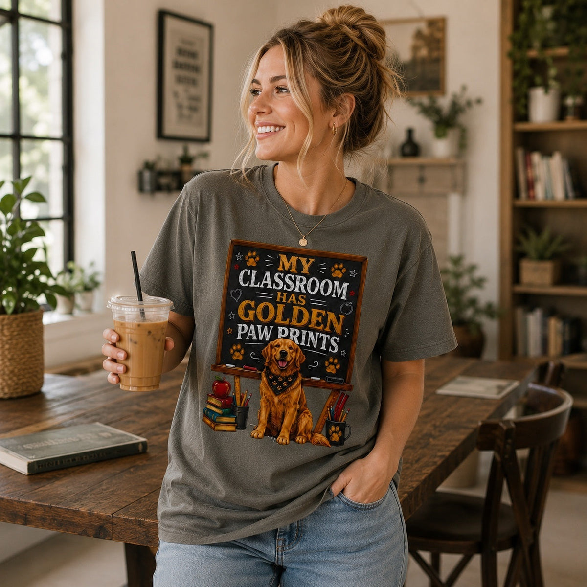 Woman wearing a pepper My Classroom Has Golden Paw Prints T-shirt with a Golden Retriever classroom design while holding iced coffee in an indoor study room.