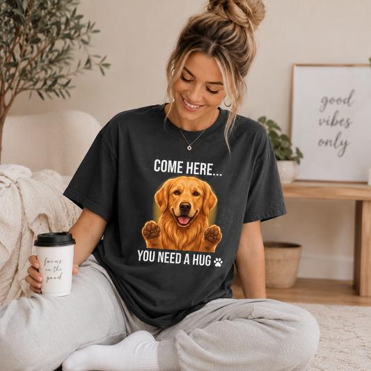 Smiling woman relaxing at home while wearing a graphite-colored Golden Retriever dog lover T-shirt with the quote “COME HERE… YOU NEED A HUG,” cozy indoor lifestyle scene with coffee cup, neutral aesthetic décor, casual loungewear, warm natural lighting, premium pet lover apparel mockup for ecommerce product listing.