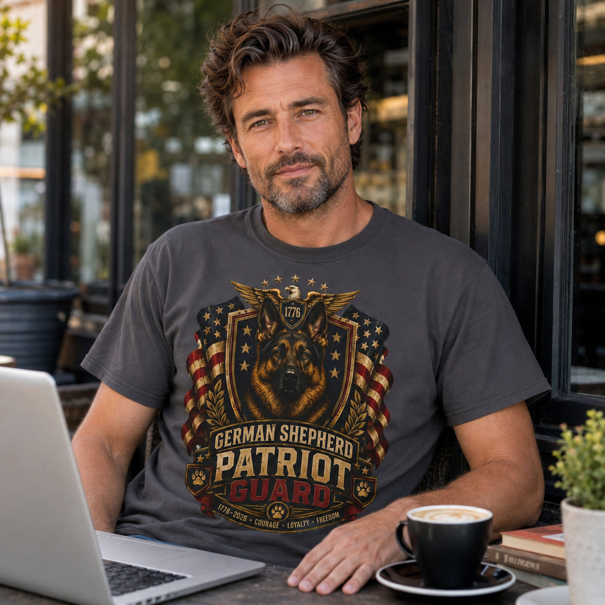 Man wearing a graphite German Shepherd Patriot Guard T-shirt with a German Shepherd patriotic crest design at a cafe with coffee and laptop.