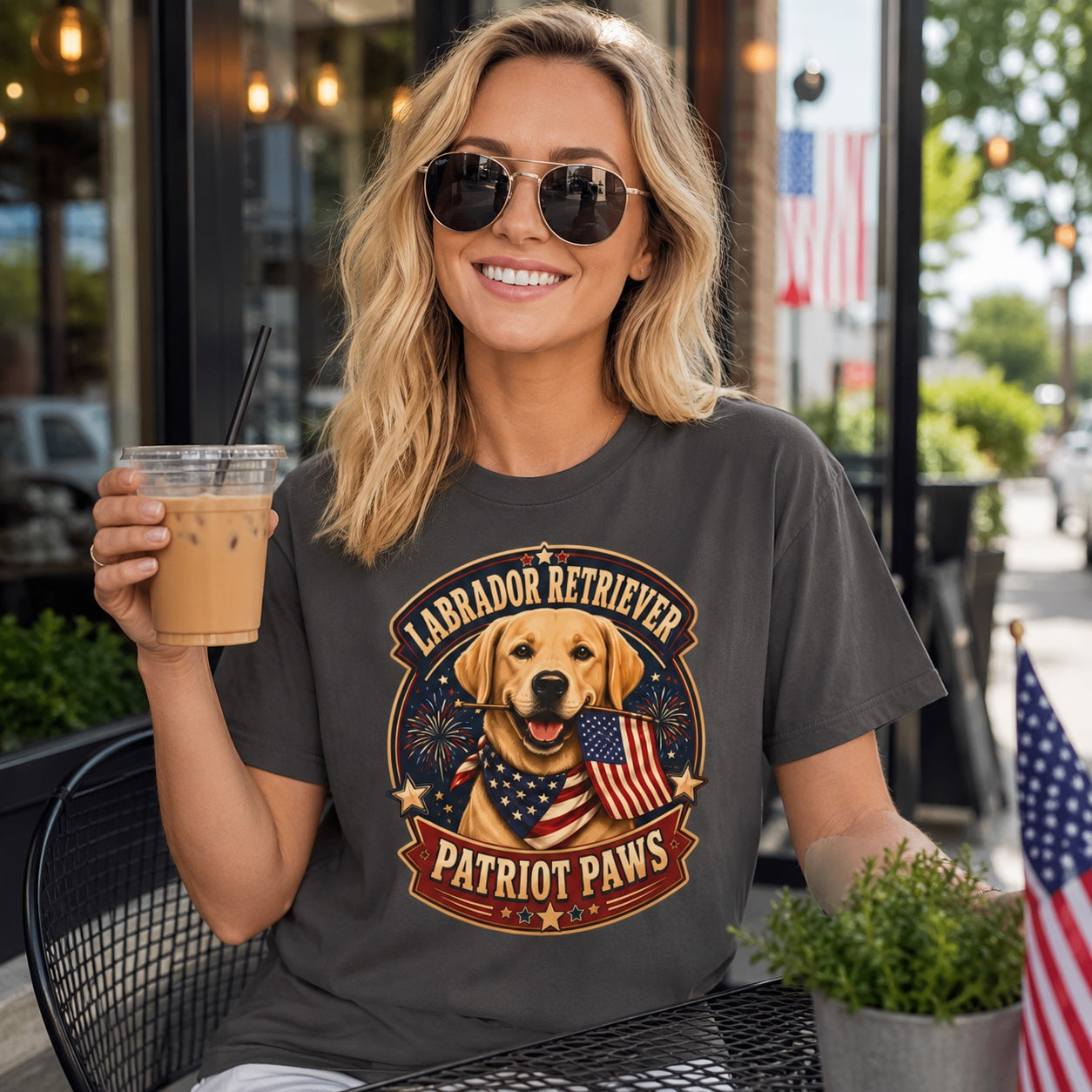 Woman in graphite patriotic dog tee wearing sunglasses, holding coffee cup next to her Labrador Retriever outdoors