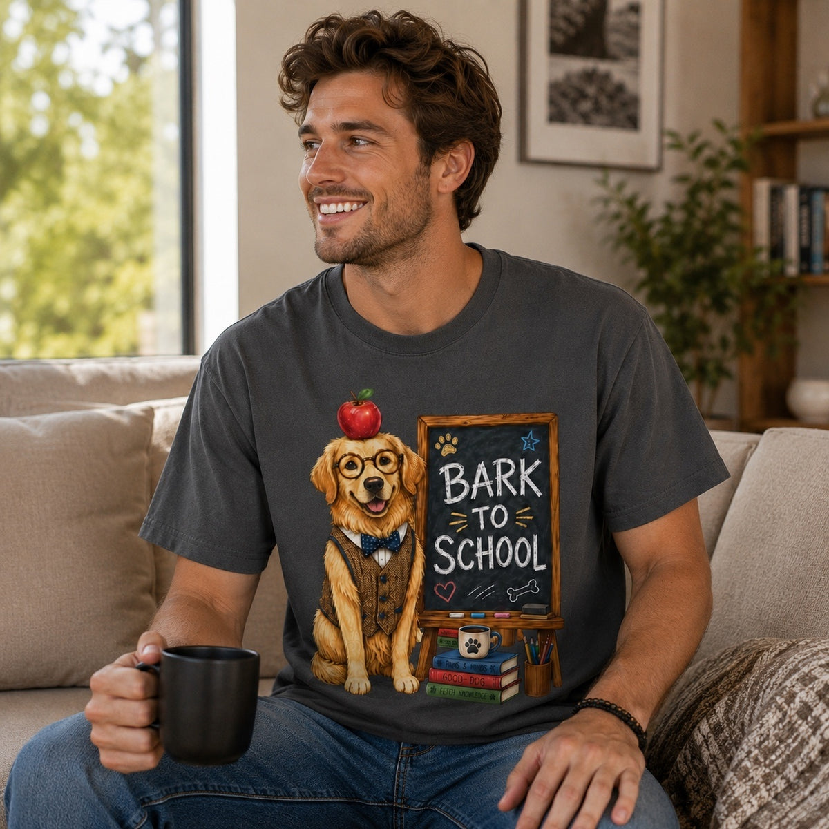 Smiling man wearing a graphite Bark To School Golden Retriever Teacher T-shirt while holding a coffee mug in a modern living room with the colorful Golden Retriever school artwork clearly visible.