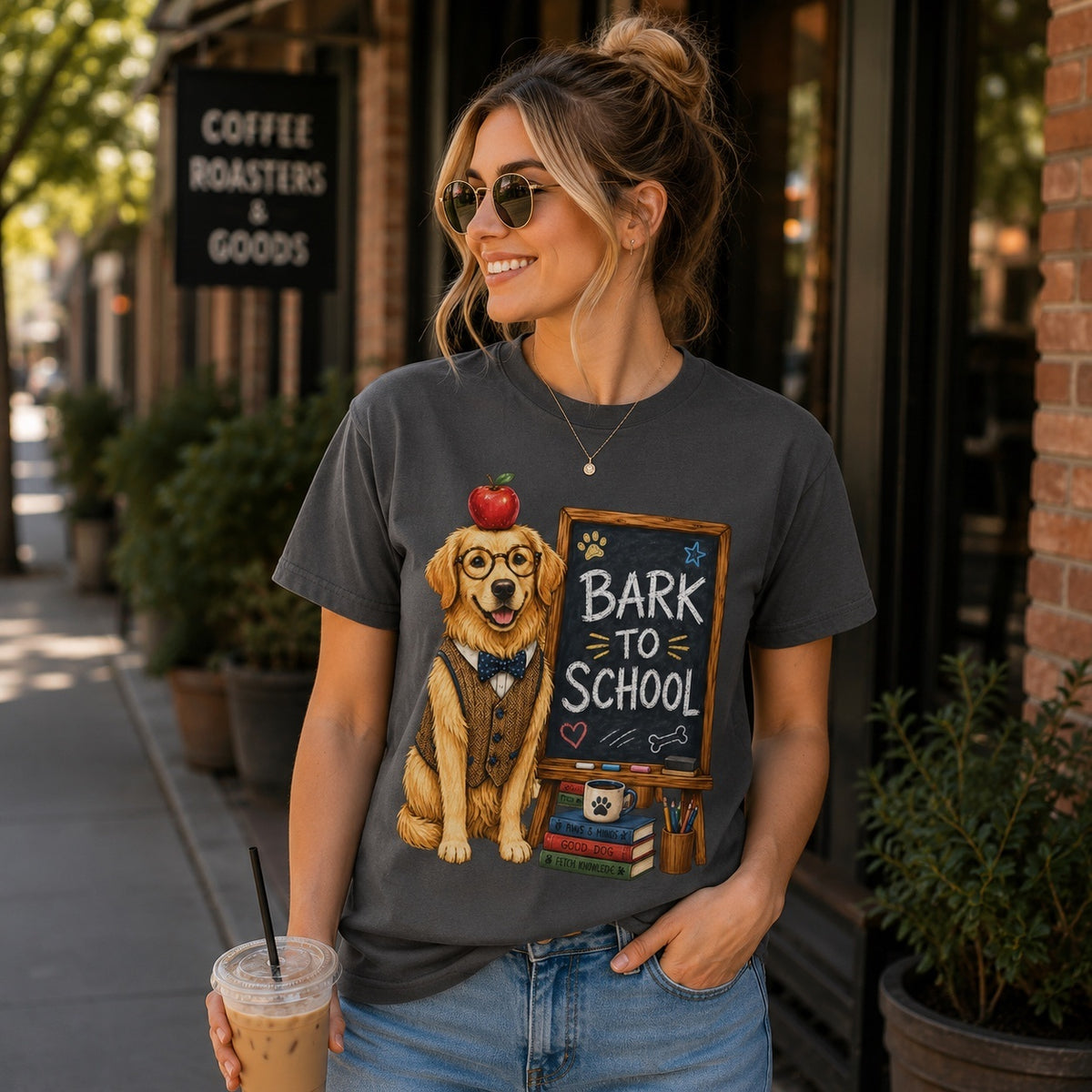 Young woman wearing a graphite Bark To School Golden Retriever teacher T-shirt while holding an iced coffee outside a coffee shop with the colorful graphic clearly visible.