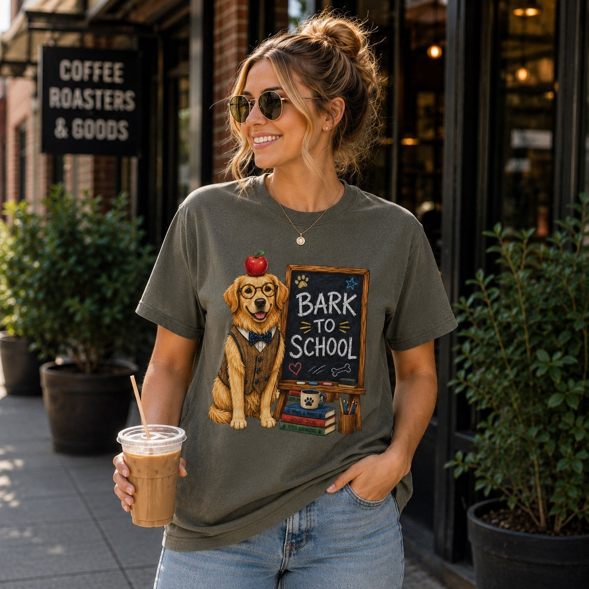 Young woman wearing a Comfort Colors Pepper “Bark to School” Golden Retriever teacher T-shirt while holding an iced coffee outside a coffee shop.