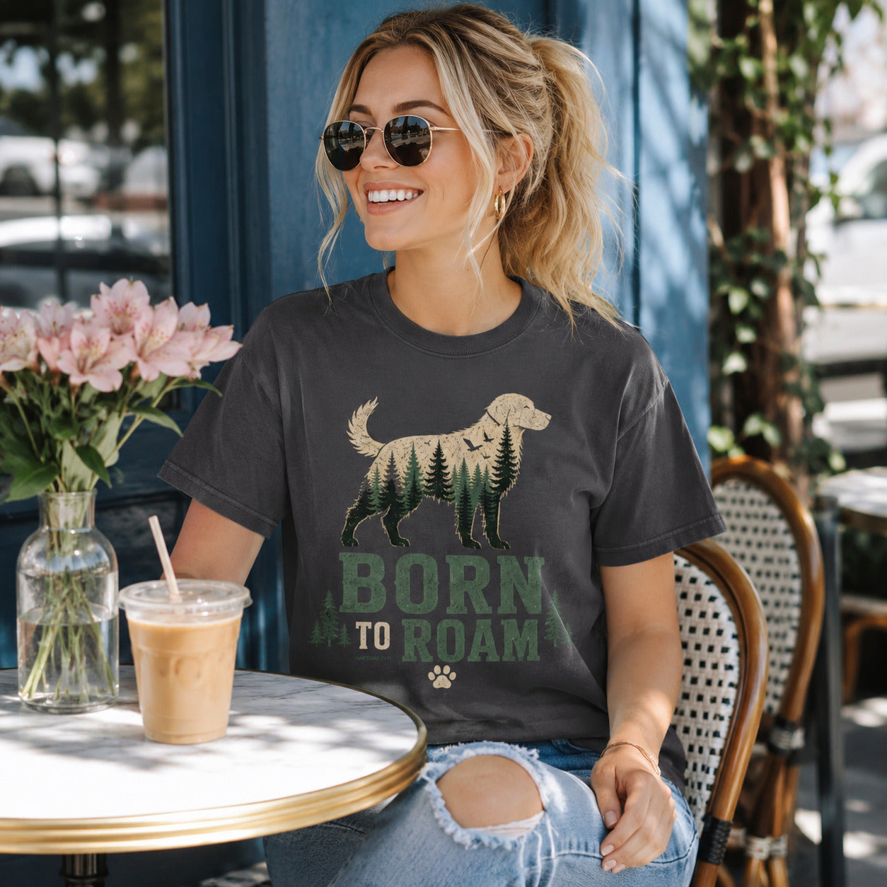 Woman sitting at an outdoor cafe table near flowers holding iced coffee, wearing graphite 'Born to Roam' dog t-shirt
