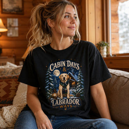 Woman wearing a black Cabin Days & Labrador Ways T-shirt with a Labrador design while sitting on a sofa inside a cozy cabin.