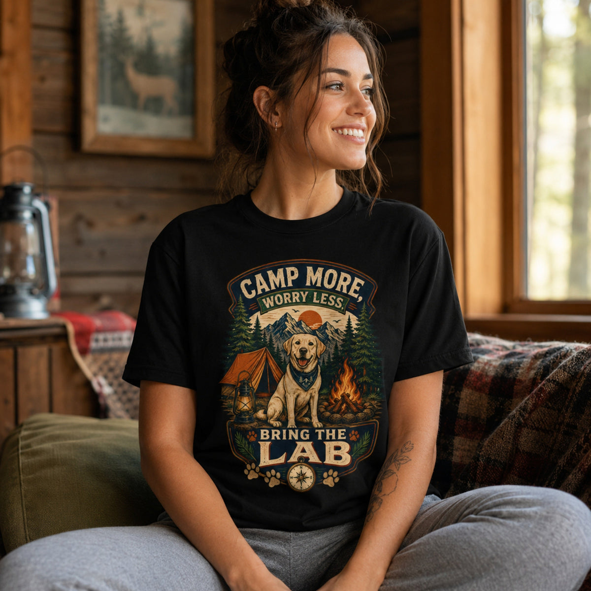 Woman wearing a black Camp More Worry Less Bring The Lab T-shirt on a cabin sofa with the Labrador campfire artwork clearly visible.