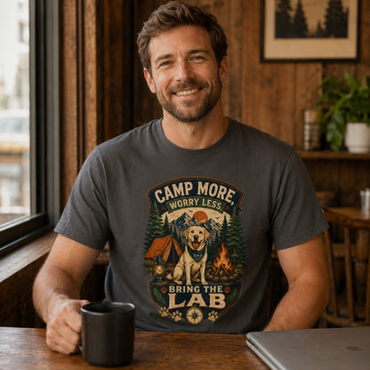 Man wearing a graphite Camp More Worry Less Bring The Lab T-shirt with coffee and laptop accessories while the Labrador camping design stays visible.