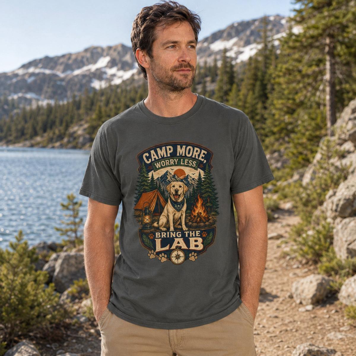 Man wearing a pepper Camp More Worry Less Bring The Lab T-shirt beside a lakeside mountain campsite with the Labrador outdoor print centered.