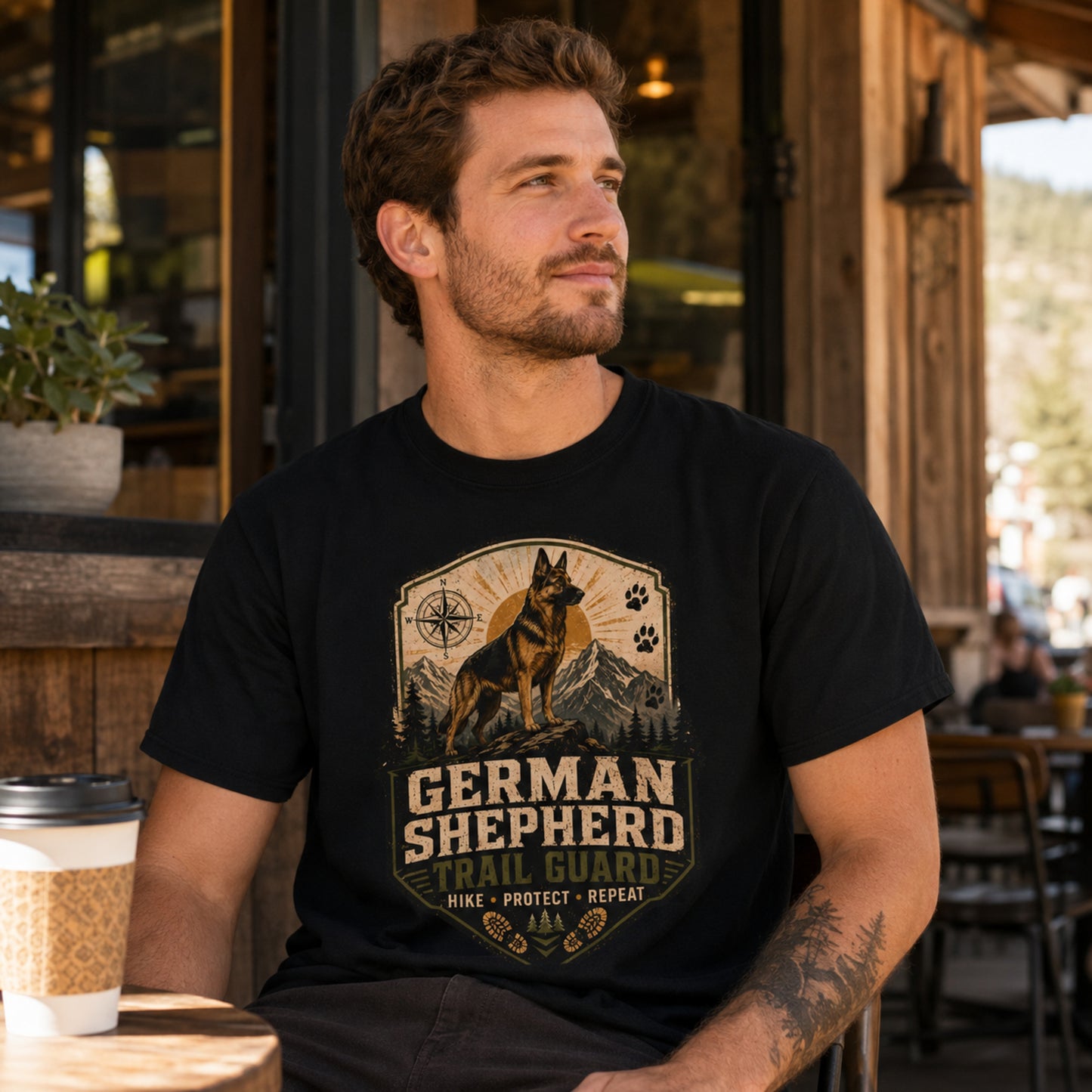 Man wearing a black German Shepherd Trail Guard T-shirt at a cafe with coffee nearby.