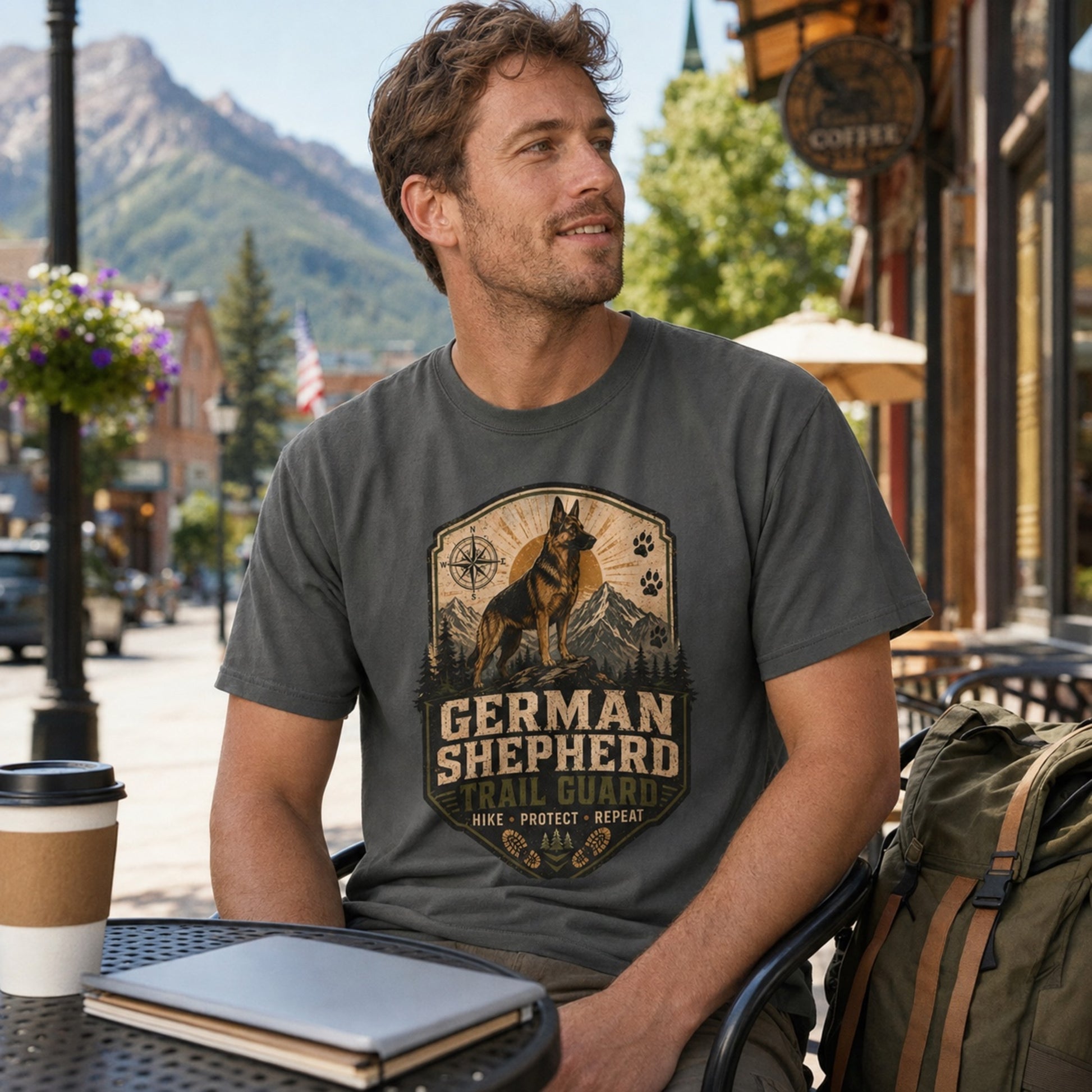 Man wearing a pepper German Shepherd Trail Guard T-shirt at an outdoor cafe with coffee, laptop, and bag nearby.