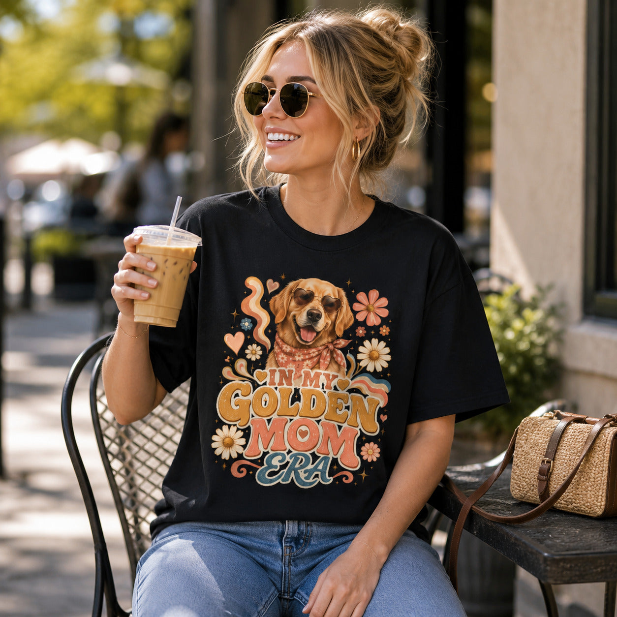 Woman sitting at an outdoor cafe holding iced coffee, wearing black 'In My Golden Mom Era' retro floral t-shirt
