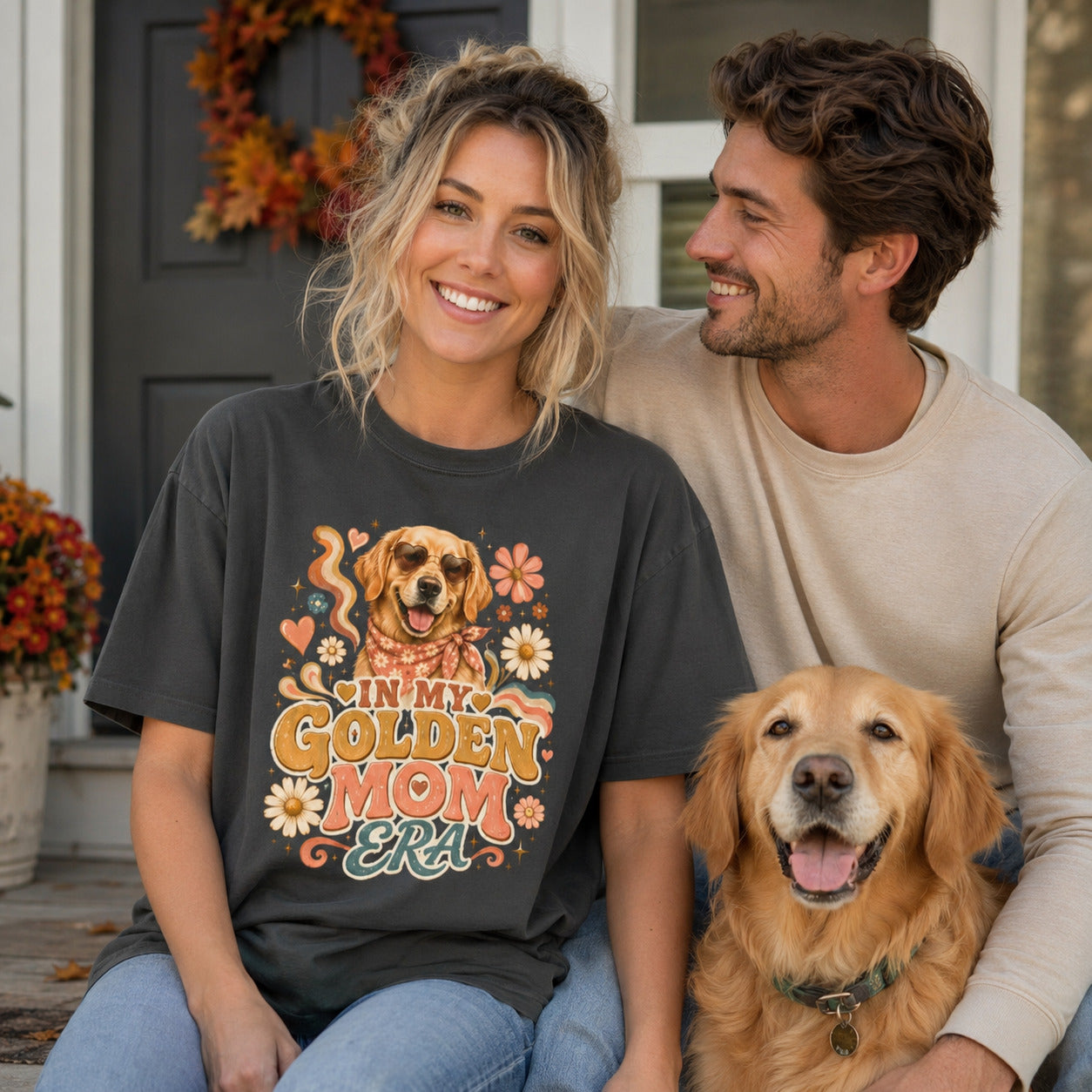 Couple sitting together with their golden retriever near a wreath-decorated door, wearing graphite 'In My Golden Mom Era' retro floral t-shirts