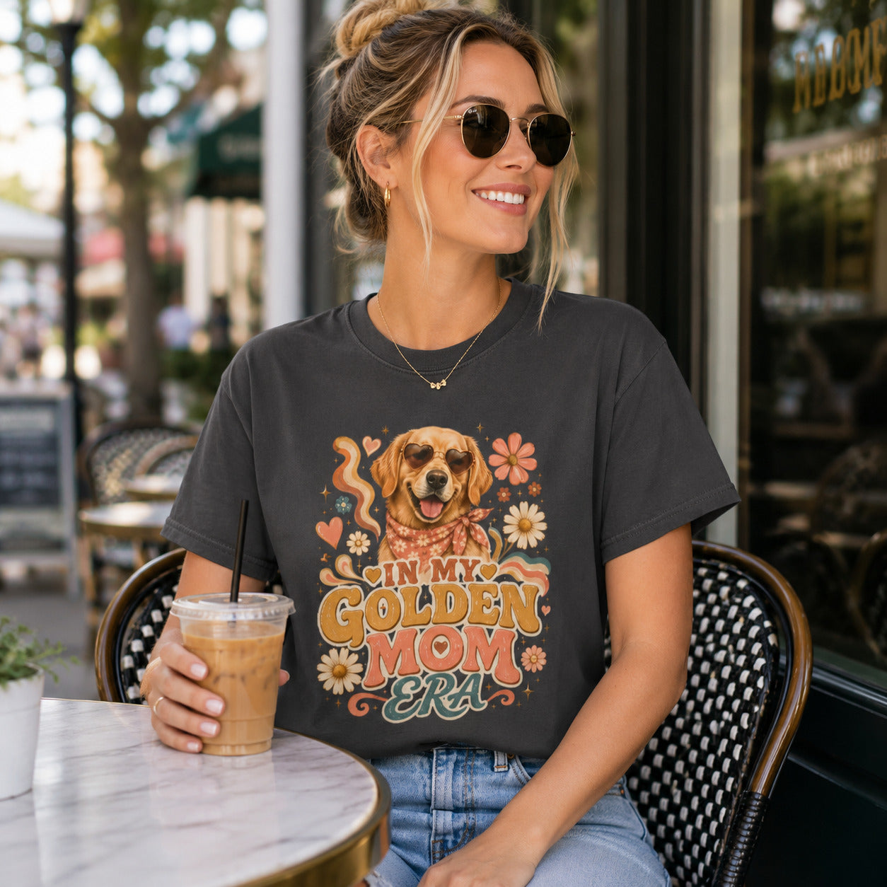 Woman sitting at an outdoor cafe with sunglasses holding iced coffee, wearing graphite 'In My Golden Mom Era' retro floral t-shirt
