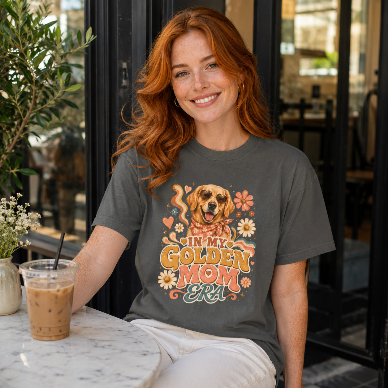 Woman sitting at an outdoor cafe holding iced coffee, wearing pepper 'In My Golden Mom Era' retro floral t-shirt