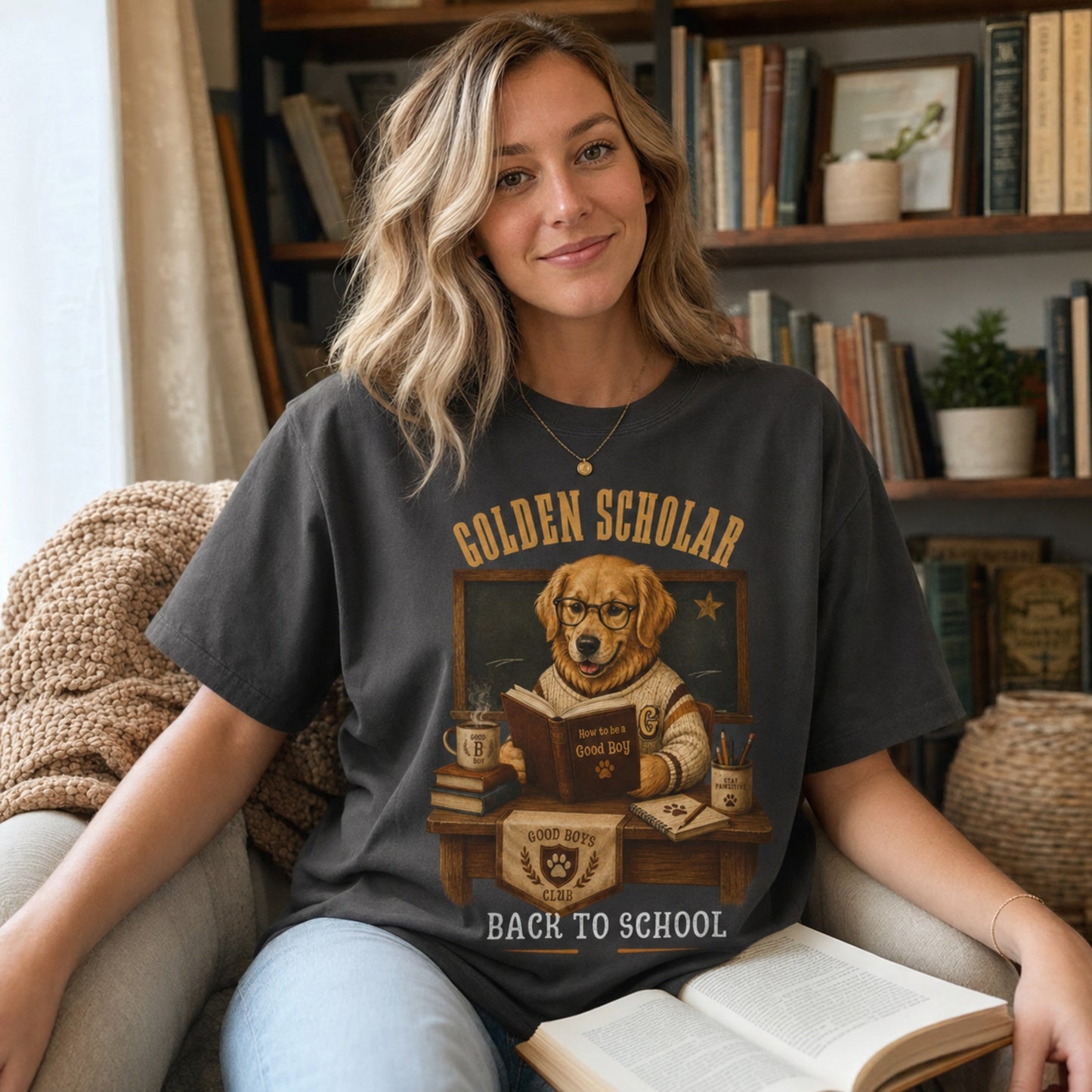 Woman wearing a graphite Golden Scholar Dog T-shirt in a reading nook with a golden retriever classroom graphic.