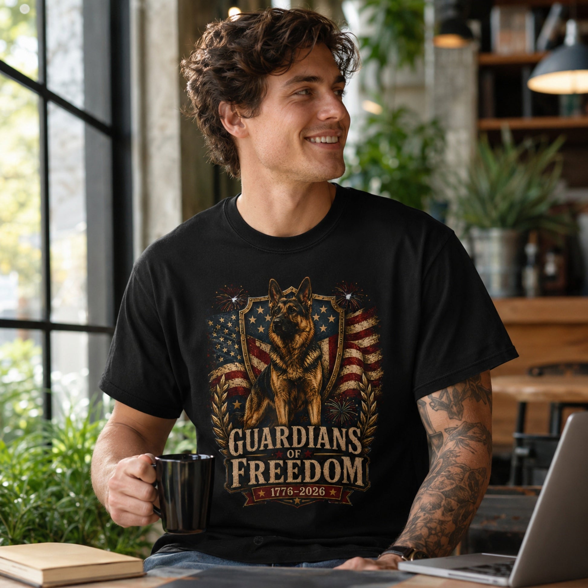 Man wearing a black Guardians of Freedom K-9 T-shirt with a German Shepherd patriotic graphic while holding a coffee mug near a laptop in a coffee shop.