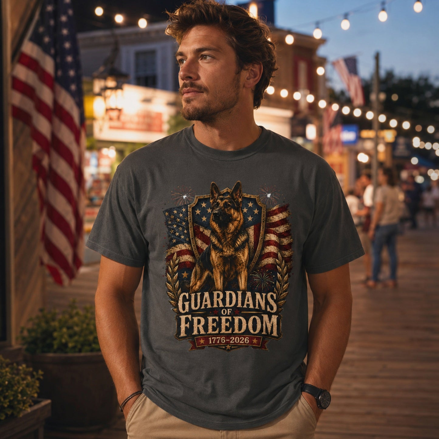 Man wearing a graphite Guardians of Freedom K-9 T-shirt with a German Shepherd patriotic graphic in an evening boardwalk scene with American flags.