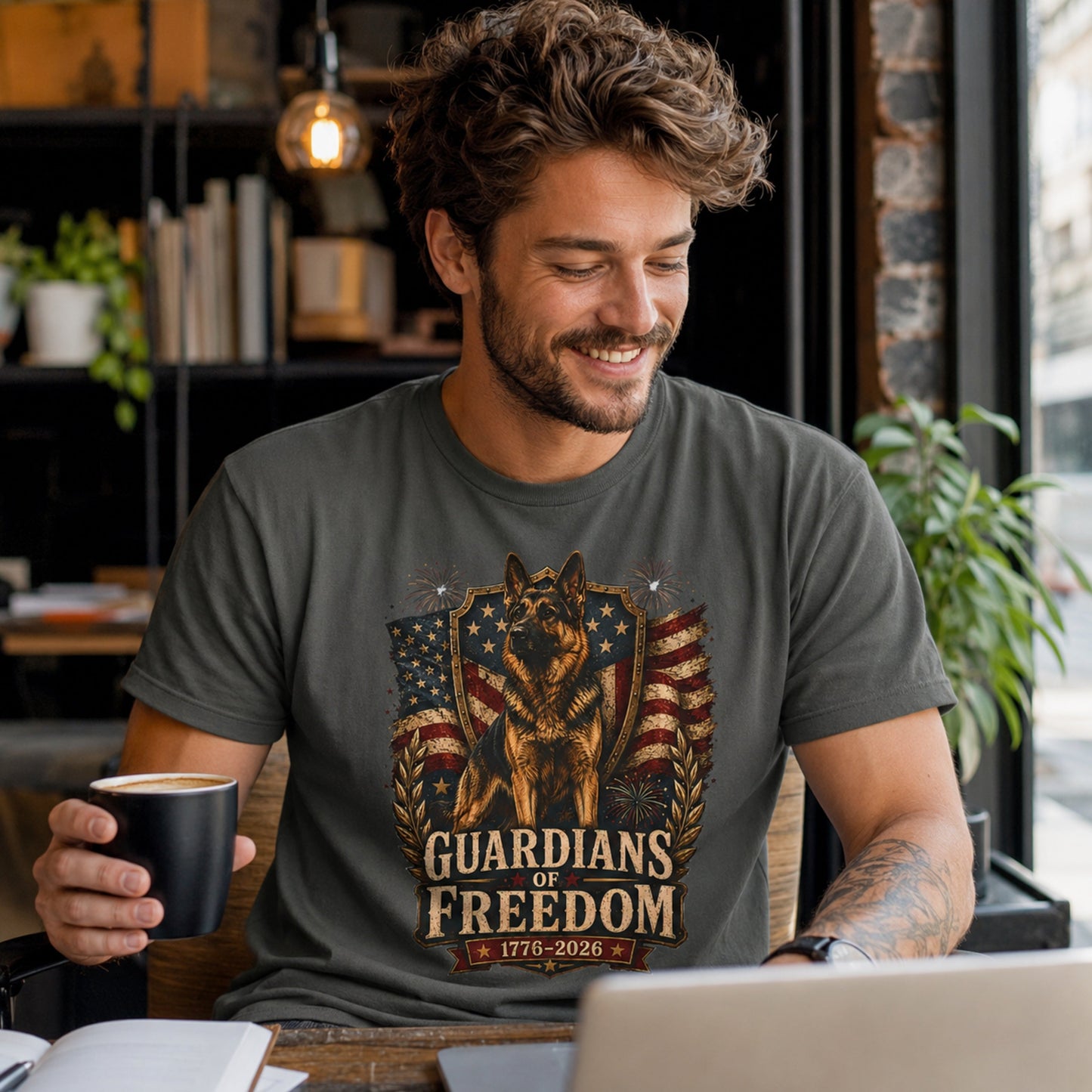 Man wearing a pepper Guardians of Freedom K-9 T-shirt with a German Shepherd patriotic graphic while holding a coffee mug near a laptop in a coffee shop.