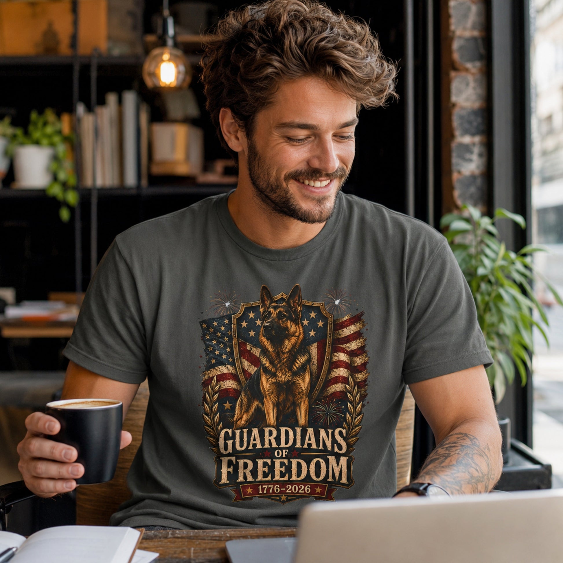 Man wearing a pepper Guardians of Freedom K-9 T-shirt with a German Shepherd patriotic graphic while holding a coffee mug near a laptop in a coffee shop.