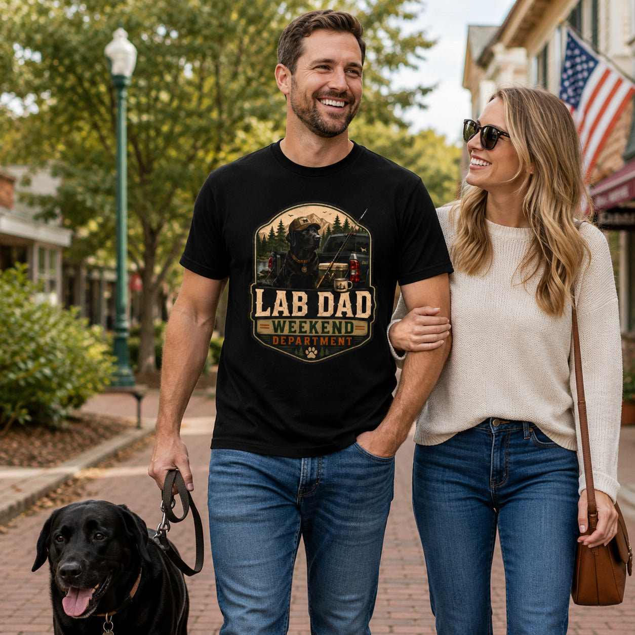 Couple walking a sidewalk with their black Labrador near an American flag, wearing black 'Lab Dad Weekend' t-shirts