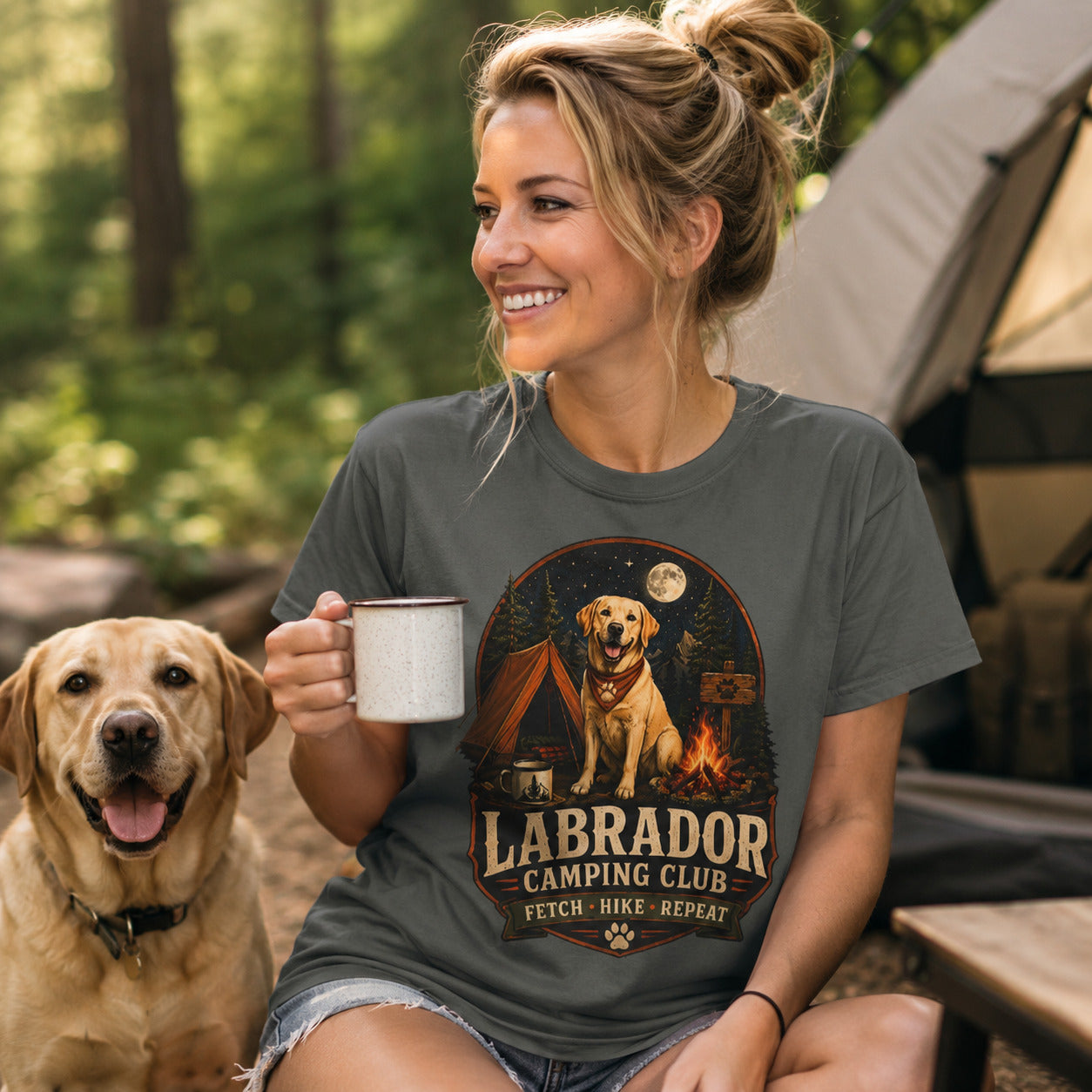 Woman wearing a pepper Labrador Camping Club Fetch Hike Repeat T-shirt holding coffee with a Labrador at a campsite.
