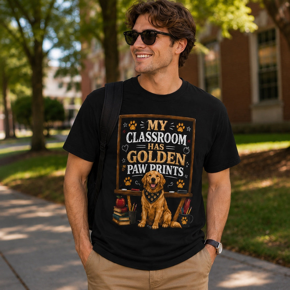 Man wearing a black My Classroom Has Golden Paw Prints T-shirt with a Golden Retriever classroom design on an outdoor school campus walkway.