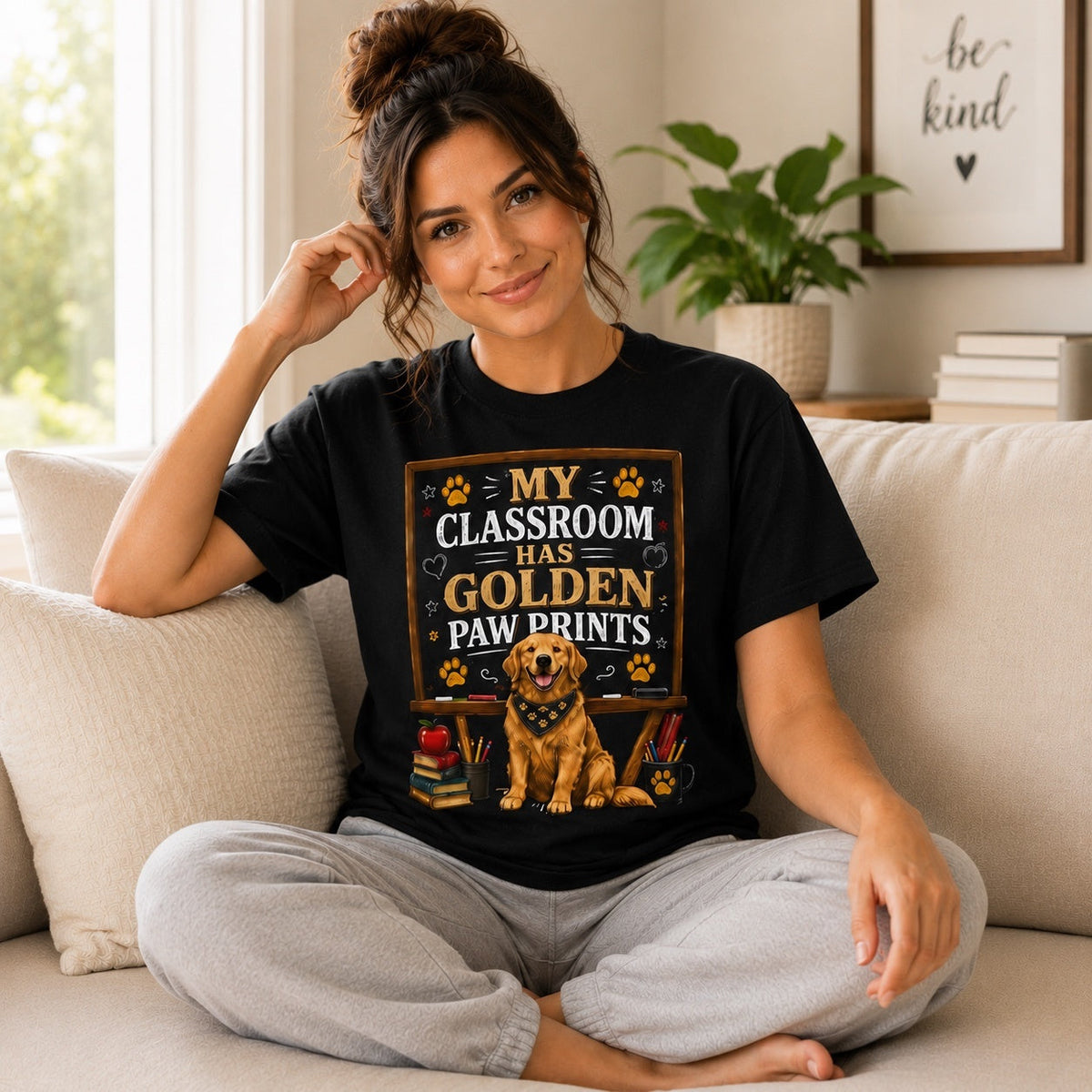 Woman wearing a black My Classroom Has Golden Paw Prints T-shirt with a Golden Retriever classroom design while sitting on a home sofa.