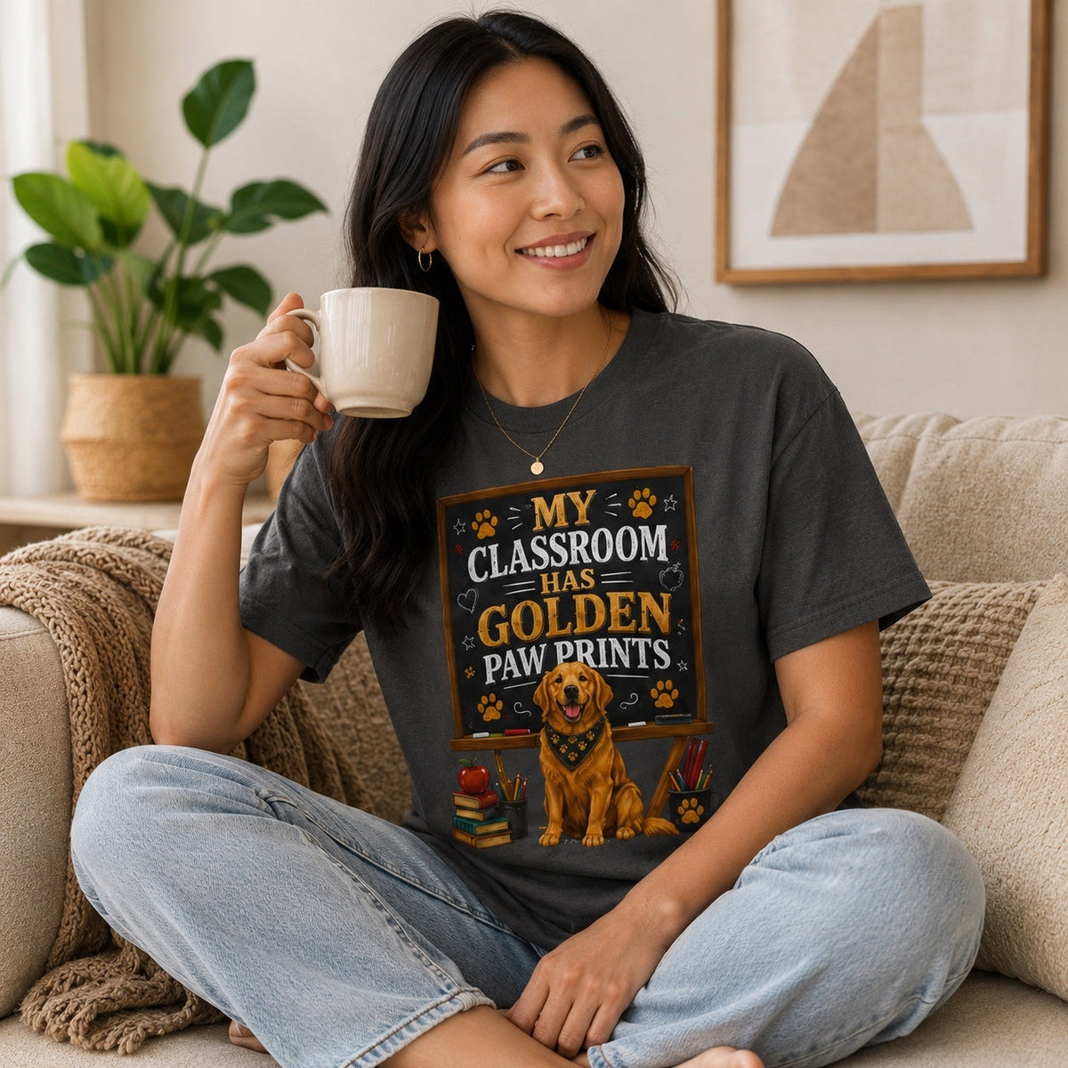 Woman wearing a graphite My Classroom Has Golden Paw Prints T-shirt with a Golden Retriever classroom design while holding coffee on a home sofa.
