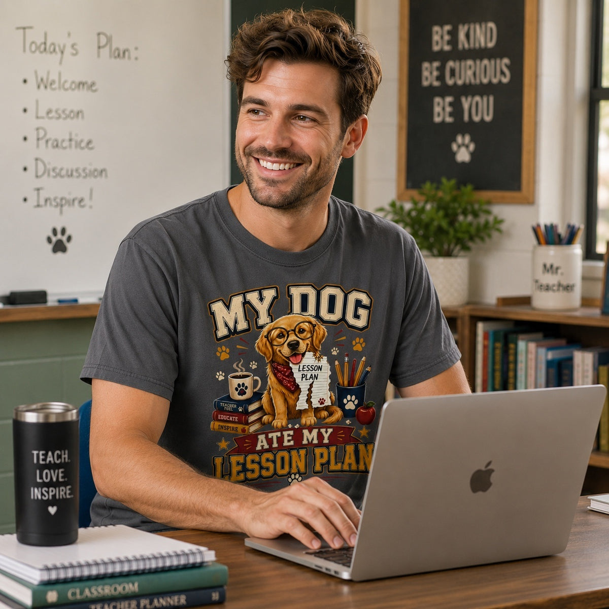 American male teacher wearing a Graphite Comfort Colors T-shirt with the “My Dog Ate My Lesson Plan” Golden Retriever graphic while working on a laptop in a classroom.