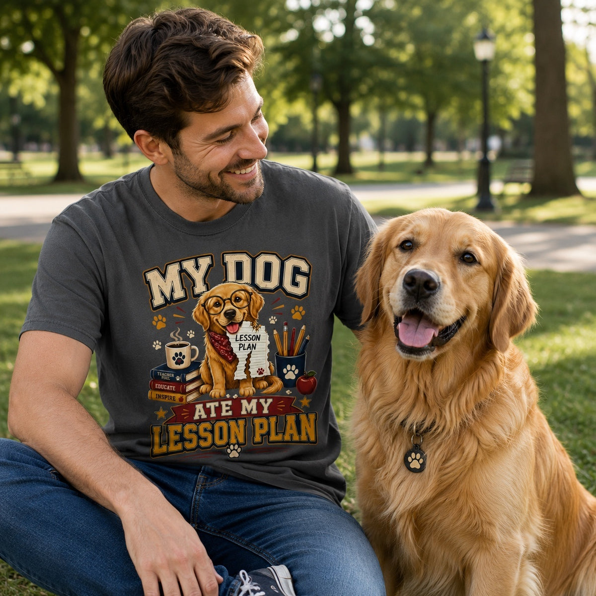 Young American man wearing a Graphite Comfort Colors T-shirt with the “My Dog Ate My Lesson Plan” Golden Retriever teacher graphic while relaxing in a park beside a Golden Retriever.