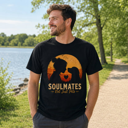 Man wearing a black Soulmates Not Just Pets T-shirt with dog silhouette sunset artwork near a lakeside park.