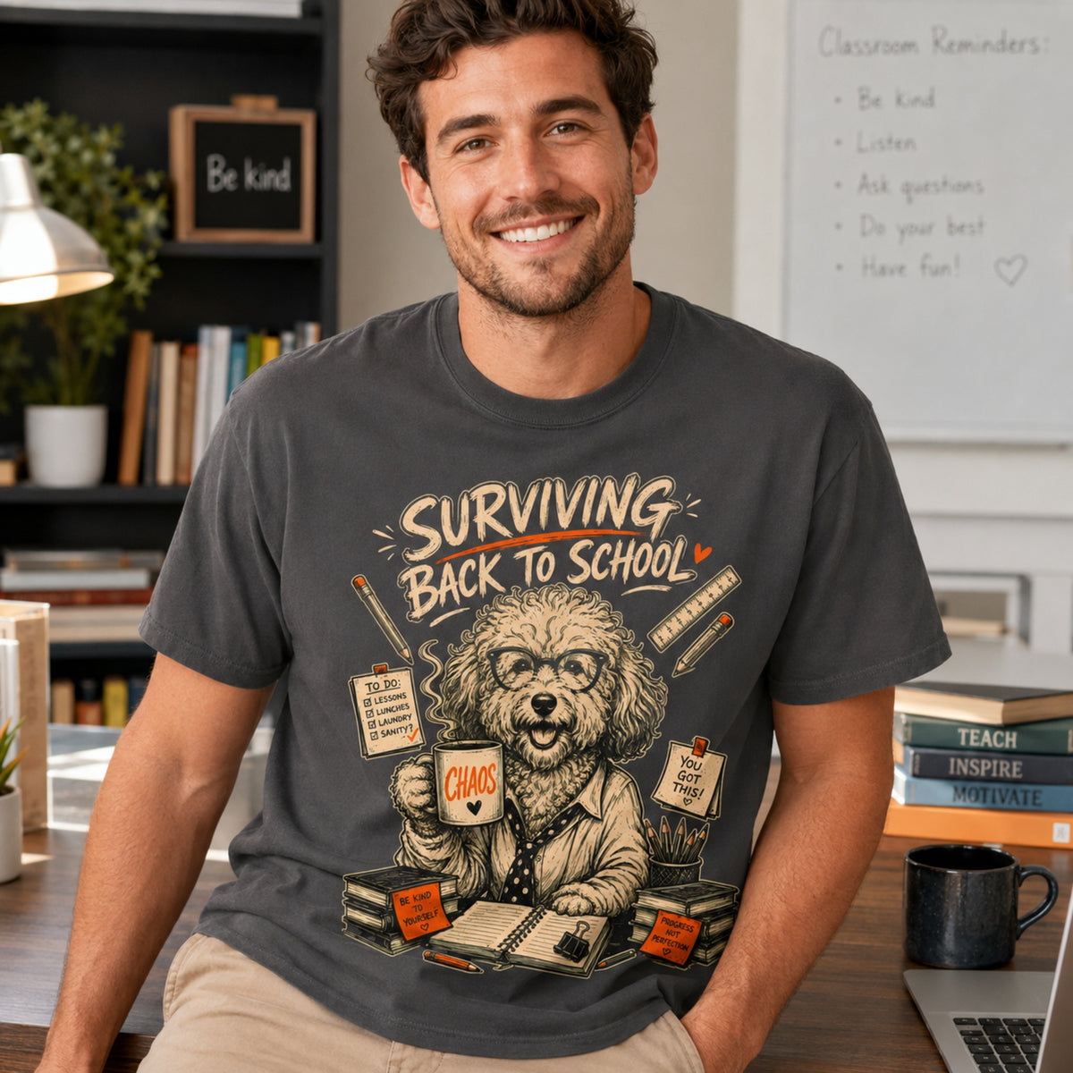 Man wearing a graphite Surviving Back to School dog teacher T-shirt with coffee, books, and a laptop.