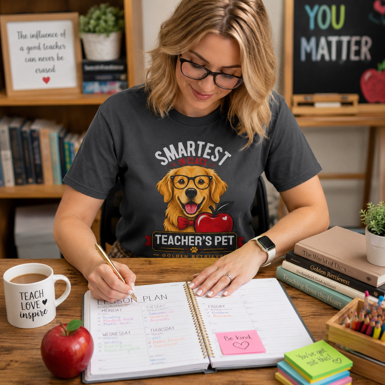 Woman writing at a desk with an apple and mug, wearing graphite 'Teacher's Pet Golden Retriever' t-shirt