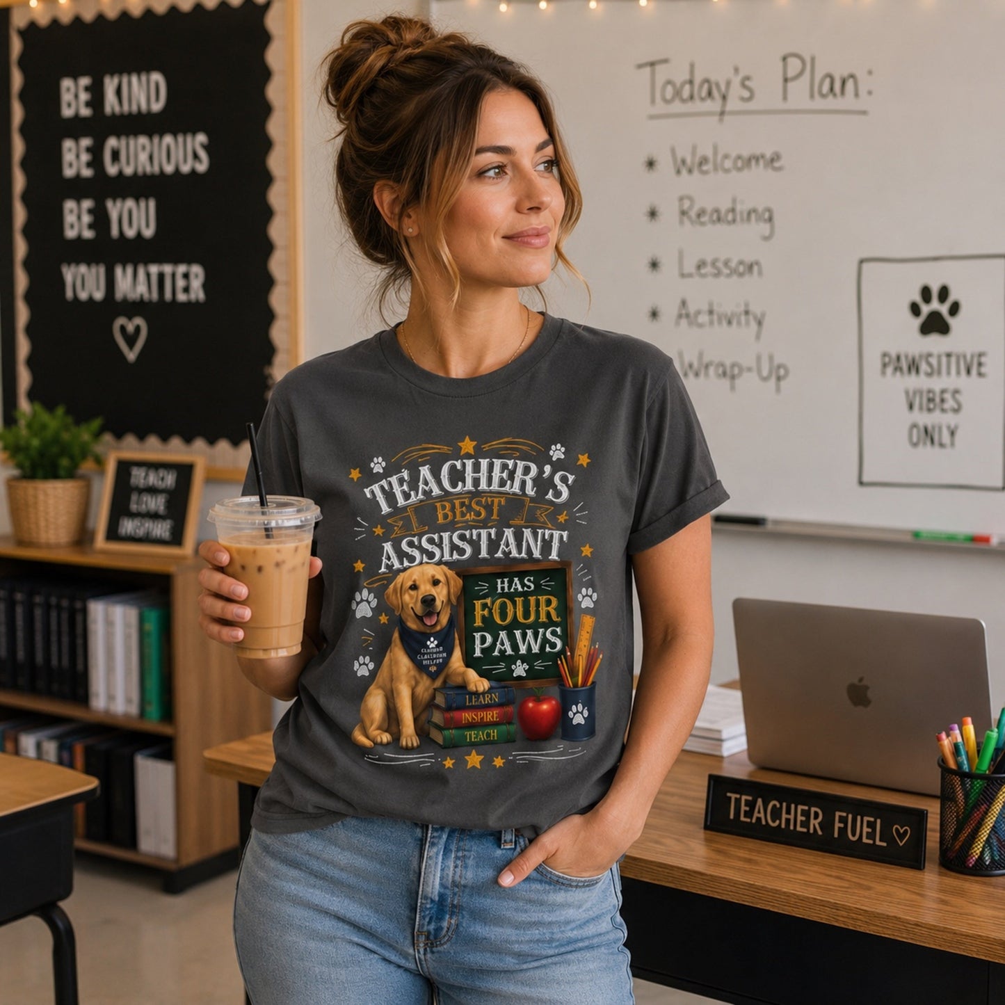 Woman wearing a graphite Teacher's Best Assistant Has Four Paws T-shirt with a Labrador classroom helper design in a classroom with iced coffee and laptop.